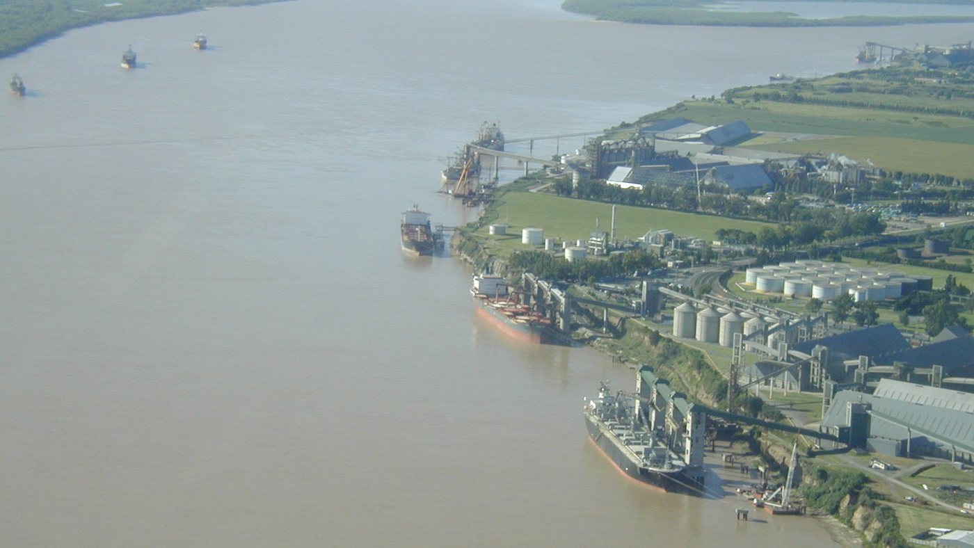 Vista aérea del río Paraná: sobre la barranca de la derecha, terminales con silos, tanques y cintas transportadoras; tres buques amarrados al muelle y otros tres navegando aguas arriba.