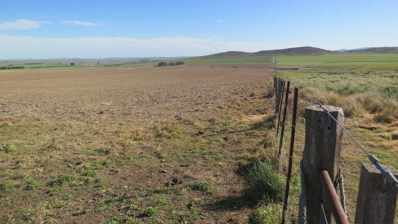 Alambrado de postes de madera entre un lote labrado y un pastizal de matas altas; al fondo, lomadas bajas y cielo despejado.