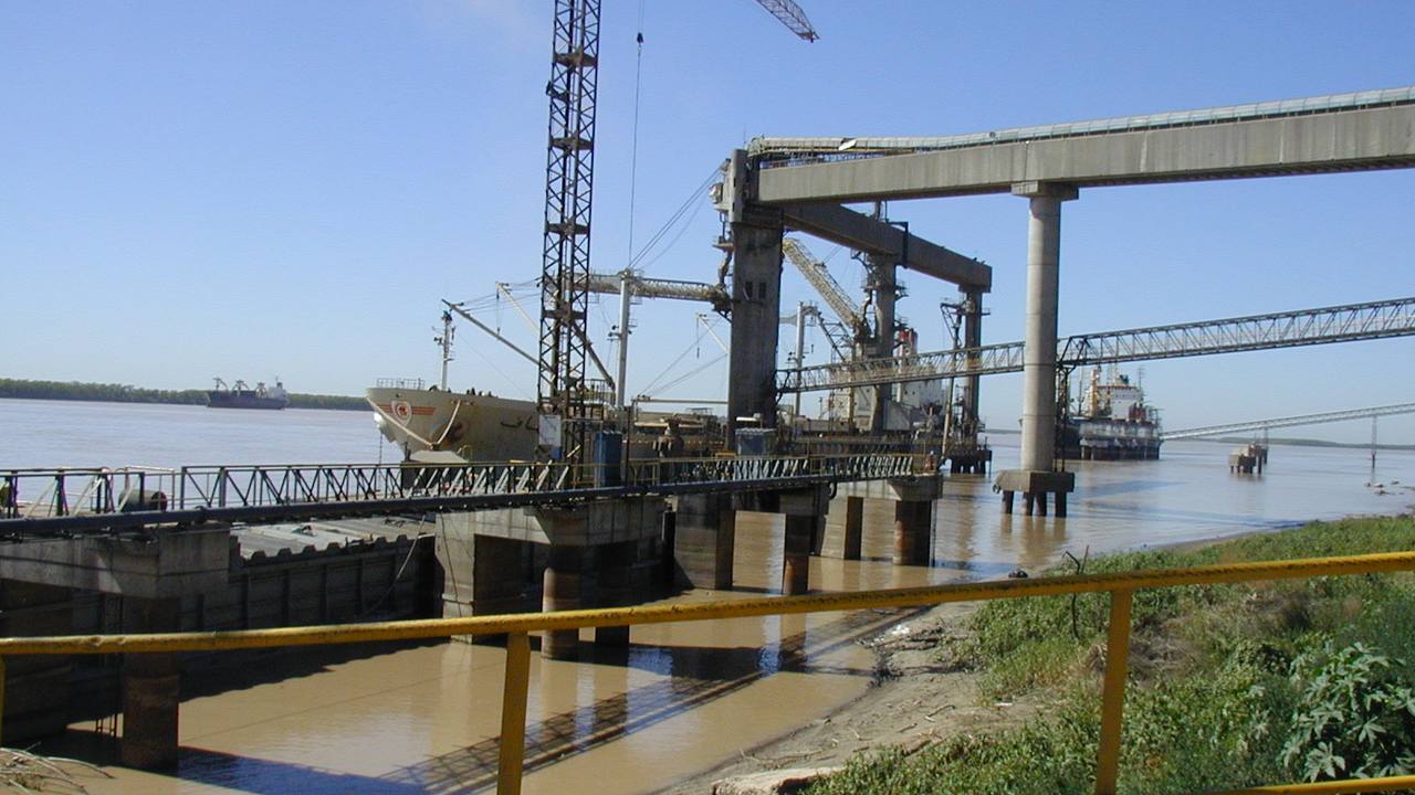 Buque granelero amarrado a un muelle de hormigón sobre el río Paraná; cintas transportadoras y torres de carga se extienden sobre el agua marrón y, en primer plano, una baranda amarilla y una barcaza.