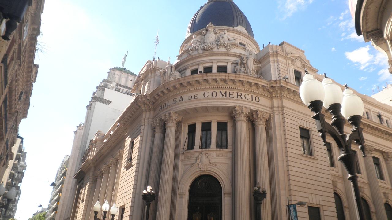 Fachada en esquina de un edificio de piedra clara, con columnas, cúpula oscura y la leyenda “BOLSA DE COMERCIO” tallada sobre el frontis.